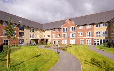 The newly built, yellow, and red bricked, three storey retirement living property set behind winding pathways which lead to a circular outdoor seating area as well the property and lovely grassed areas.