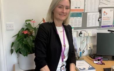 A white woman with blonde hair, wearing a dark suit jacket and white top, with a pink lanyard round her neck, sat at a desk in an office and smiling at the camera.