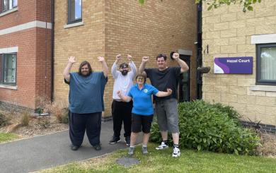 A group of five individuals standing in front of the brick facade of the Hazel Court building. They are all smiling and making hand gestures above their heads to mark their celebration