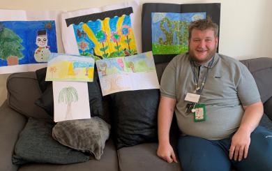 A white man with fair hair in his 30s, sat on a sofa in a lounge, surrounded by his handpainted artwork, smiling at the camera.