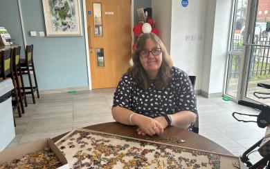 A white woman with shoulder-length brown hair and glasses, sat at a round table in front of a half-completed jigsaw showing giraffes, and smiling at the camera.