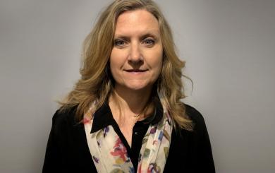 A white woman with long blonde hair, wearing a dark blouse and floral scarf, smiling at the camera.