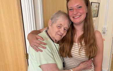 An older woman with short hair and a light green top hugging a young woman with long brown hair wearing a striped dress. They are standing indoors, smiling, with a wooden door and a painting in the background