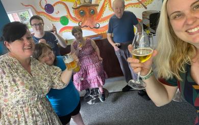 A group of six white men and women in their 30s, 40s and 50s, standing in a room decorated like a pub, each holding a drink and smiling to the camera.