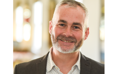 Headshot of a man wearing a shirt with a pattern of small dots and a dark suit jacket, smiling at the camera. The background is an indoor space with soft lighting, suggesting a professional setting