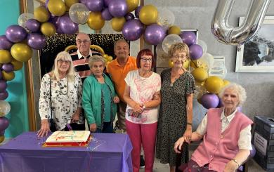 A group of people in their 70s and 80s smiling at the camera, surrounded by a purple and gold balloon arch, with a cake in front of them.