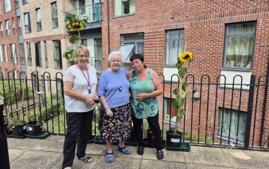 Three women standing on a patio at Rose Manor, proudly displaying two tall sunflowers in pots beside them. The women, including resident Kath Knox (centre), are smiling, with the red-brick exterior of Rose Manor and windows of the building visible in the background.