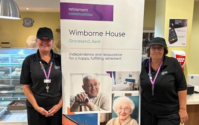 Staff members of Wimborne House's catering team, standing in the restaurant wearing black uniforms, smiling beside a promotional banner for the retirement community in Gravesend, Kent. The banner features images of residents and the building.