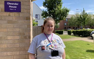 A woman who appears to have Down syndrome, wearing a grey t-shirt and a purple work lanyard, standing in front of a building with greenery in the background