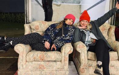 Two young women sit on floral sofas, presumably donated from a TV set. Both wear cozy clothes and beanie hats, smiling as the woman on the right strikes a playful pose, framing her face with one hand while flailing her other arm out.