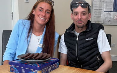 A young woman wearing a sky blue blazer with long auburn hair smiles alongside a young man with a goatee-style moustache and beard, wearing a black gilet and cap. Both have sunglasses resting on their heads, and there is a plate of chocolate biscuits in front of them.