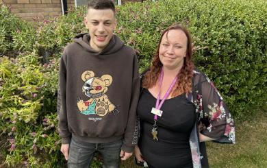  A young man in a brown hoodie smiles next to a woman with mid-length auburn hair wearing a sheer kimono-style cardigan over a black dress and a purple lanyard. They stand in front of a Buddleja bush, with the man slightly taller.