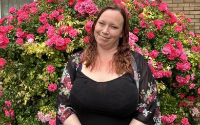 Young woman with mid-length hair in front of a bush full of pink flowers, clasping her hands together and smiling for the camera.