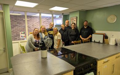  A group of eight white men and women in their 20s, 30s and 40s, standing together and smiling at the camera. They're stood in a kitchen with freshly-painted green walls. In front of them is a kitchen island with two cookers and a pot of utensils. They're all smiling at the camera.