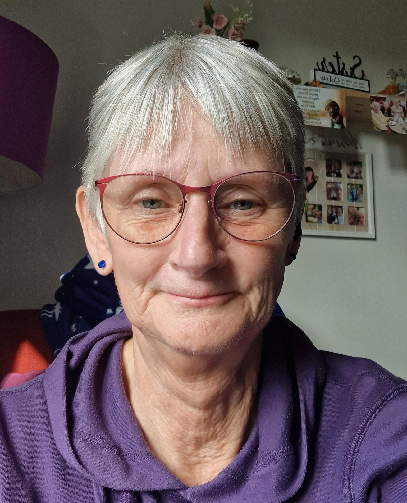 A white woman in her 70s, sat on a sofa in her flat, smiling at the camera.