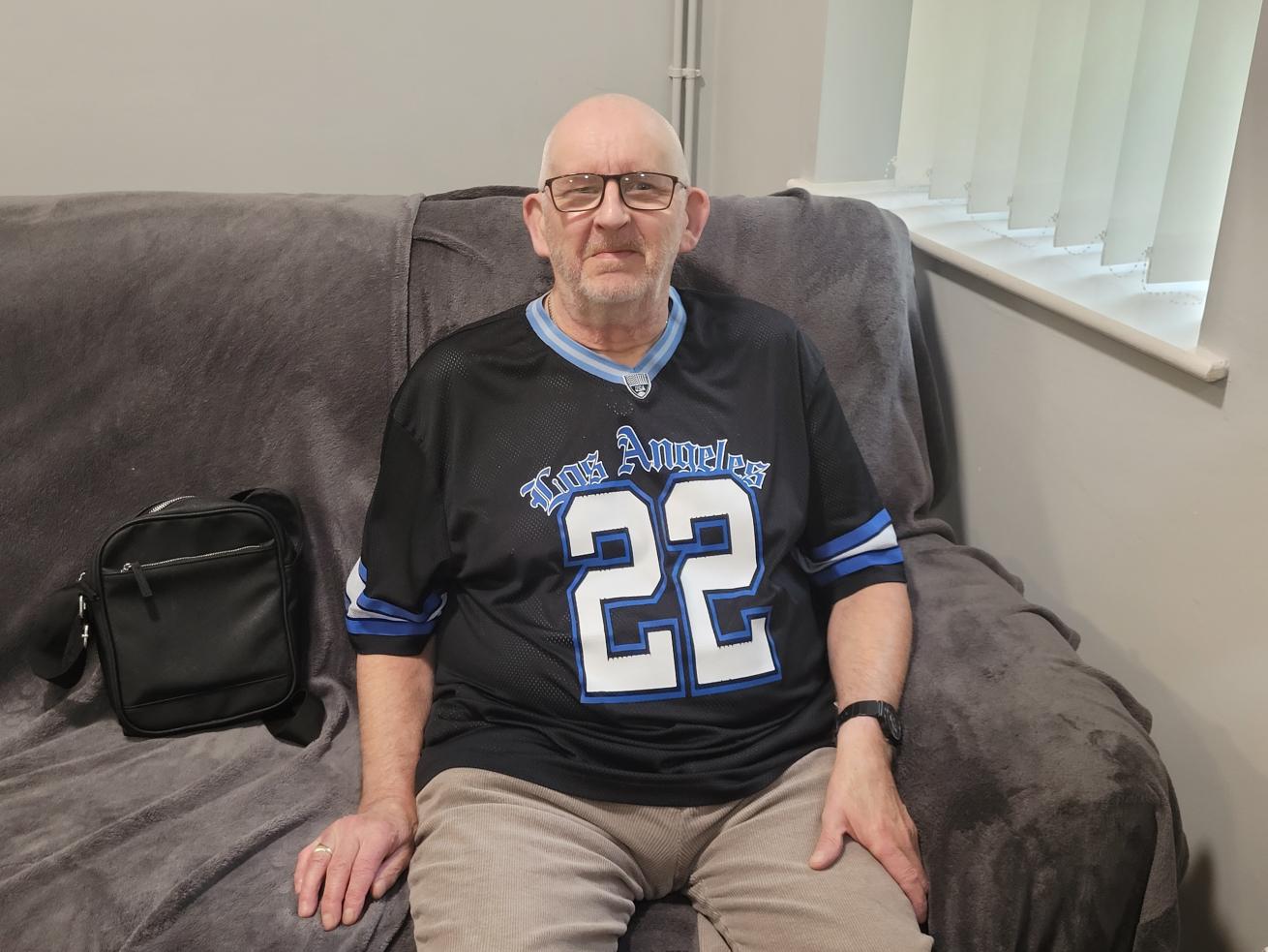  An older white man wearing glasses and a dark Los Angeles sports shirt, sitting on a cream sofa and smiling at the camera.