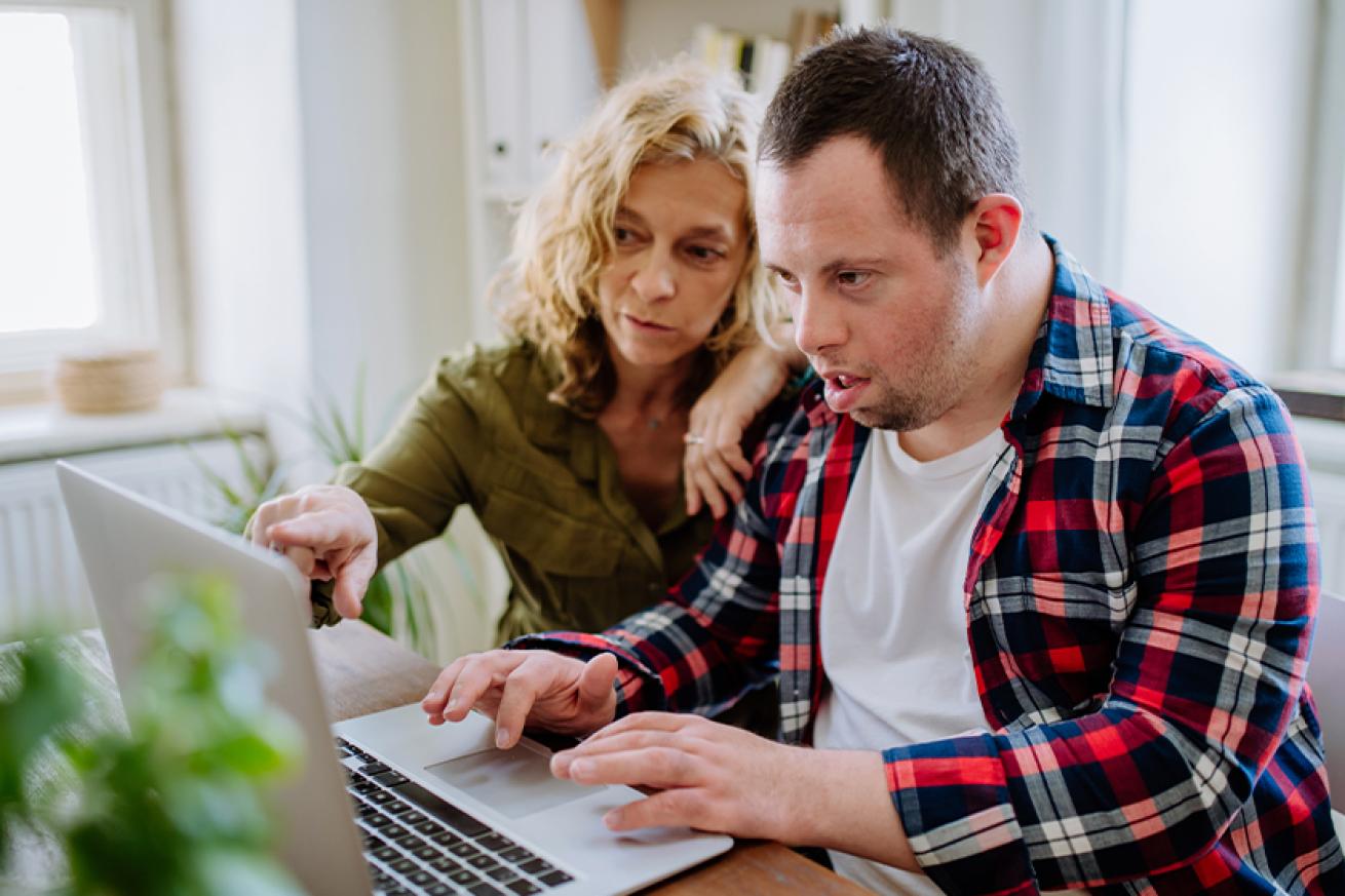 Stock photo of a young man with down syndrome wearing a red chequered shirt using a laprop on a wooden table with the help of an older woman wearing a green shirt