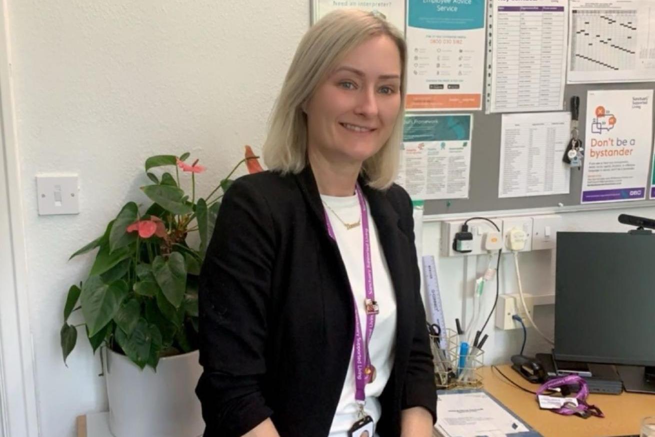 A white woman with blonde hair, wearing a dark suit jacket and white top, with a pink lanyard round her neck, sat at a desk in an office and smiling at the camera.