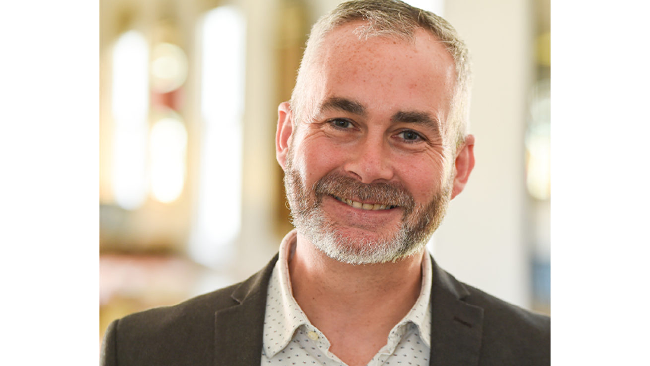 Headshot of a man wearing a shirt with a pattern of small dots and a dark suit jacket, smiling at the camera. The background is an indoor space with soft lighting, suggesting a professional setting