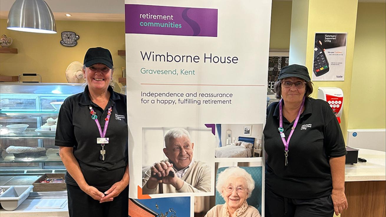 Staff members of Wimborne House's catering team, standing in the restaurant wearing black uniforms, smiling beside a promotional banner for the retirement community in Gravesend, Kent. The banner features images of residents and the building.