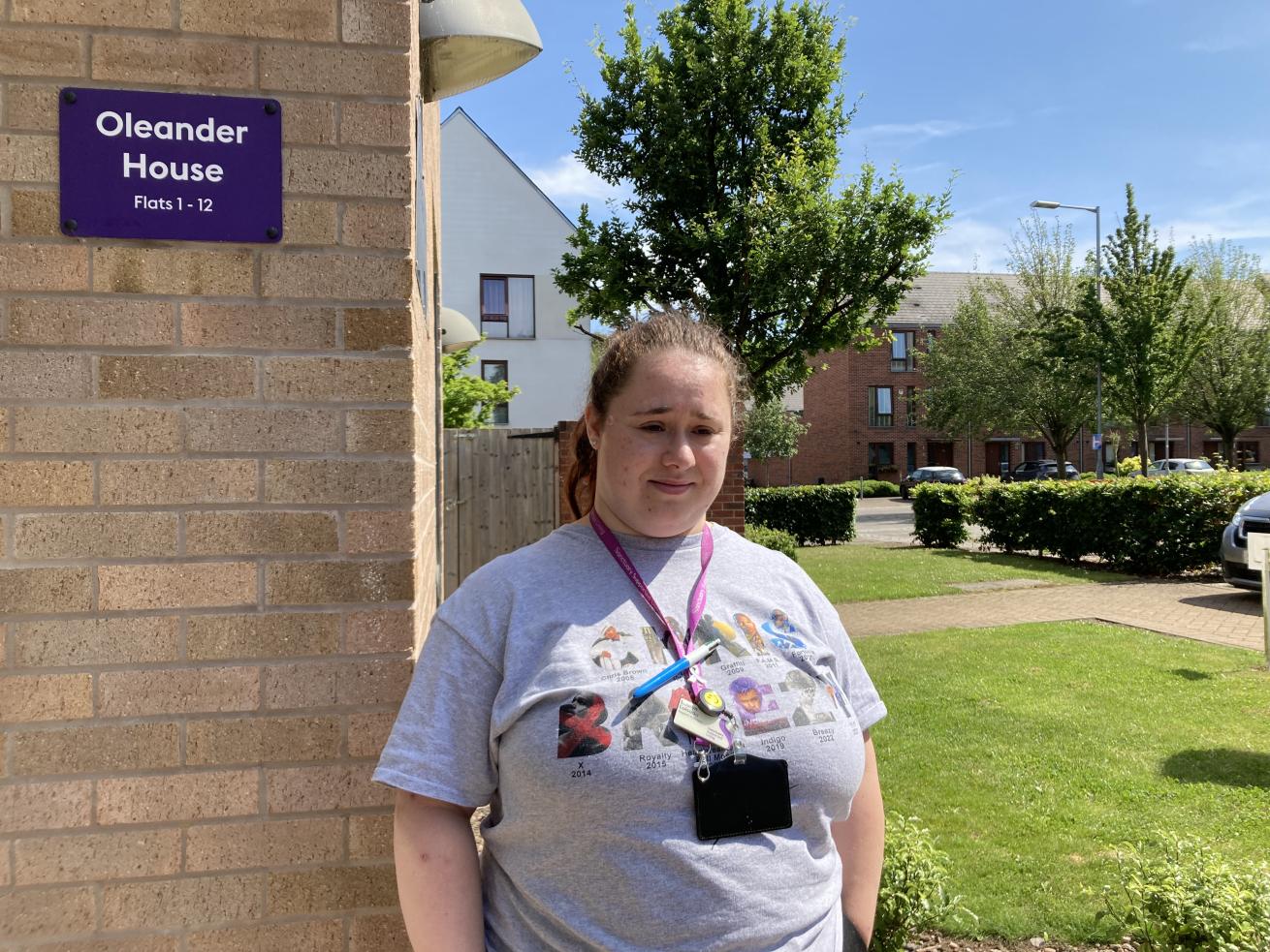 A woman who appears to have Down syndrome, wearing a grey t-shirt and a purple work lanyard, standing in front of a building with greenery in the background