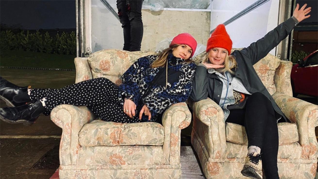 Two young women sit on floral sofas, presumably donated from a TV set. Both wear cozy clothes and beanie hats, smiling as the woman on the right strikes a playful pose, framing her face with one hand while flailing her other arm out.