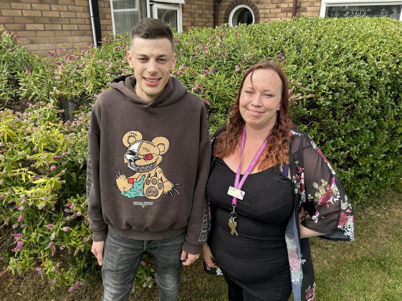  A young man in a brown hoodie smiles next to a woman with mid-length auburn hair wearing a sheer kimono-style cardigan over a black dress and a purple lanyard. They stand in front of a Buddleja bush, with the man slightly taller.