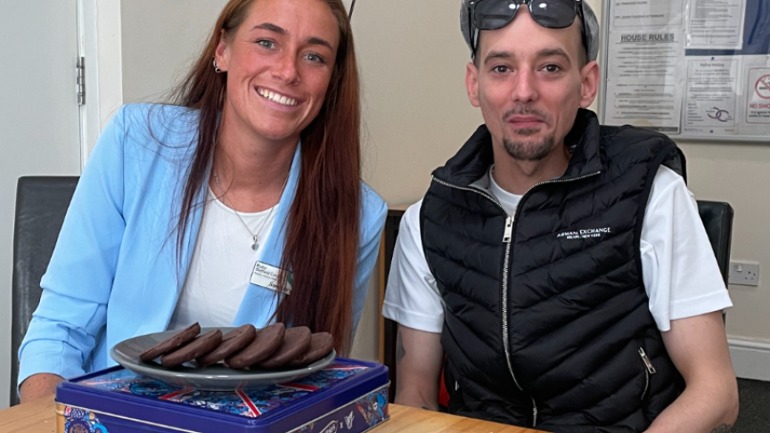 A young woman wearing a sky blue blazer with long auburn hair smiles alongside a young man with a goatee-style moustache and beard, wearing a black gilet and cap. Both have sunglasses resting on their heads, and there is a plate of chocolate biscuits in front of them.