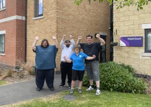 A group of five individuals standing in front of the brick facade of the Hazel Court building. They are all smiling and making hand gestures above their heads to mark their celebration