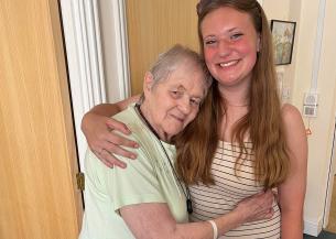 An older woman with short hair and a light green top hugging a young woman with long brown hair wearing a striped dress. They are standing indoors, smiling, with a wooden door and a painting in the background