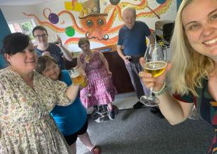 A group of six white men and women in their 30s, 40s and 50s, standing in a room decorated like a pub, each holding a drink and smiling to the camera.