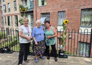 Three women standing on a patio at Rose Manor, proudly displaying two tall sunflowers in pots beside them. The women, including resident Kath Knox (centre), are smiling, with the red-brick exterior of Rose Manor and windows of the building visible in the background.