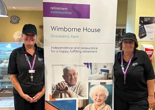 Staff members of Wimborne House's catering team, standing in the restaurant wearing black uniforms, smiling beside a promotional banner for the retirement community in Gravesend, Kent. The banner features images of residents and the building.