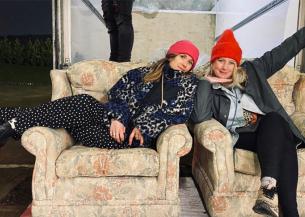 Two young women sit on floral sofas, presumably donated from a TV set. Both wear cozy clothes and beanie hats, smiling as the woman on the right strikes a playful pose, framing her face with one hand while flailing her other arm out.