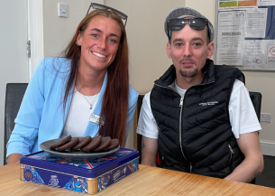 A young woman wearing a sky blue blazer with long auburn hair smiles alongside a young man with a goatee-style moustache and beard, wearing a black gilet and cap. Both have sunglasses resting on their heads, and there is a plate of chocolate biscuits in front of them.