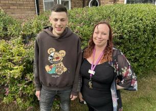  A young man in a brown hoodie smiles next to a woman with mid-length auburn hair wearing a sheer kimono-style cardigan over a black dress and a purple lanyard. They stand in front of a Buddleja bush, with the man slightly taller.