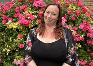 Young woman with mid-length hair in front of a bush full of pink flowers, clasping her hands together and smiling for the camera.