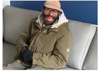 A black man wearing a green coat and hat, sat on a grey sofa against a blue wall, smiling at the camera